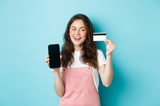 Ragazza allegra in abiti estivi che mostra lo schermo dello smartphone e la carta di credito in plastica, pagando online, facendo shopping, in piedi su sfondo blu