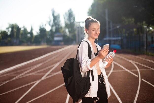 Ragazza allegra in auricolari che usa felicemente il cellulare con una bottiglia d'acqua in mano e uno zaino in spalla mentre trascorre del tempo sull'ippodromo dello stadio