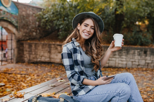 Ragazza allegra in blue jeans che si siede sulla tavola di legno e che gode del buon giorno di autunno. La signora abbastanza caucasica con i capelli castano chiaro beve il latte in cortile la mattina di ottobre.