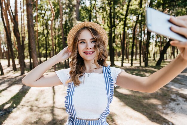 Ragazza allegra in cappello che tiene telefono e posa con un sorriso timido. Signora bianca vaga agghiacciante nella foresta nel fine settimana.