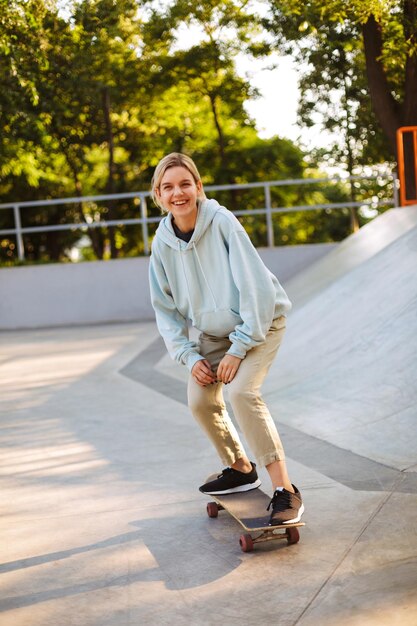 Ragazza allegra in felpa con cappuccio che guarda felicemente a porte chiuse mentre prova a fare skateboard al moderno skatepark