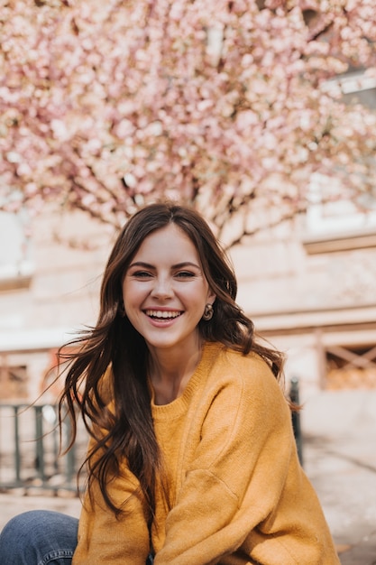 Ragazza allegra in maglione di cachemire ride sullo sfondo di sakura sbocciante. Ritratto di donna in felpa con cappuccio gialla in città in primavera