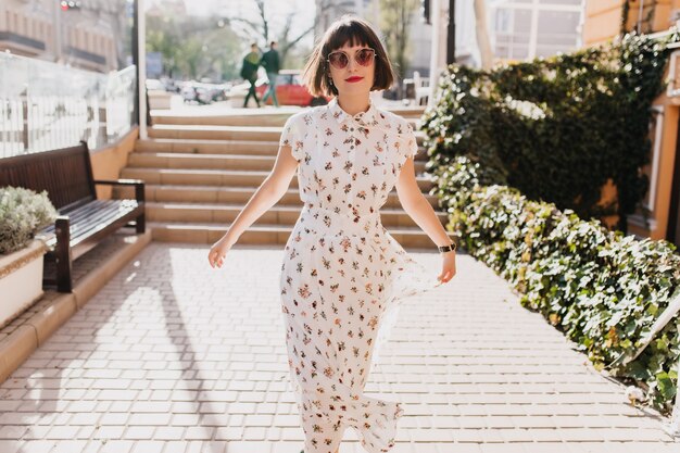 Ragazza allegra in vestito bianco dell'annata che cammina per strada in primavera. Spettacolare donna europea con i capelli corti che si rilassano in estate.