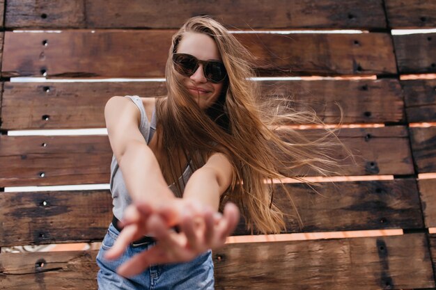 Ragazza allegra in vetri neri divertendosi sulla parete di legno. Colpo esterno del modello caucasico attraente in jeans agghiacciante in primavera.