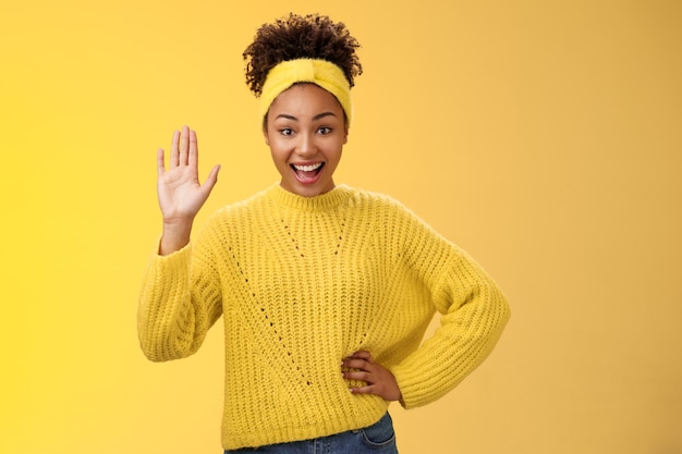 Ragazza alzando la mano volentieri disposti a partecipare essere candidato dire ciao salutando agitando gesto di saluto palmo sorridente ampiamente sentirsi felice con gioia accogliere gli amici invitando festa vicino a sfondo giallo porta.