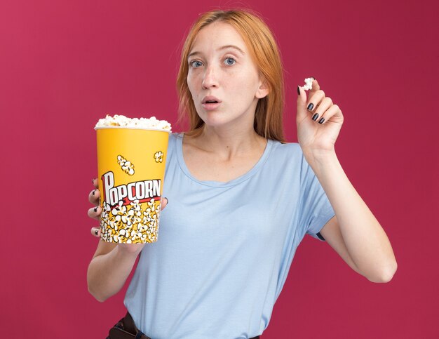 Ragazza ansiosa dello zenzero giovane rossa con le lentiggini che tengono il secchio del popcorn sul colore rosa