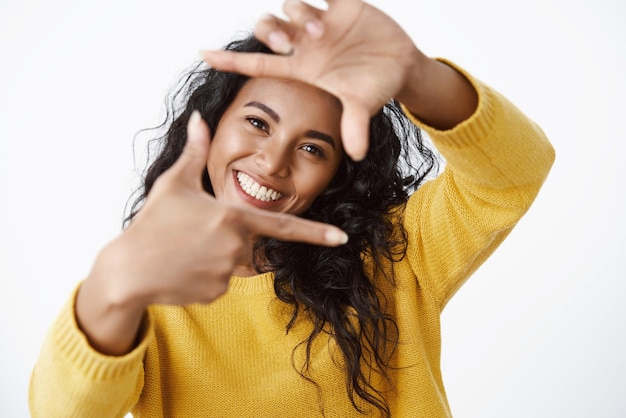 Ragazza artista alla ricerca di ispirazione nei dintorni che mostrano il gesto dei fotogrammi e guardando attraverso la forma della forma dell'obiettivo della fotocamera e sorridente ampiamente in piedi su sfondo bianco entusiasta