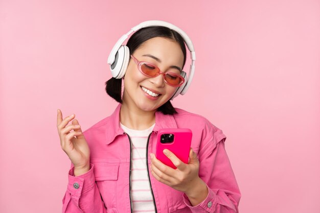 Ragazza asiatica alla moda che balla con la musica d'ascolto dello smartphone nelle cuffie sull'app del telefono cellulare che sorride e che ride in posa su sfondo rosa