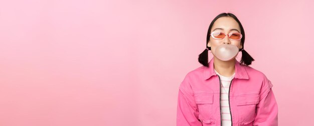 Ragazza asiatica alla moda che soffia gomma da masticare bolla di gomma da masticare indossando occhiali da sole in posa su sfondo rosa