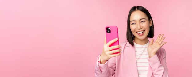 Ragazza asiatica alla moda della donna di affari in vestito che prende selfie sulla chat video dello smartphone con l'app del telefono cellulare che posa contro lo sfondo rosa dello studio