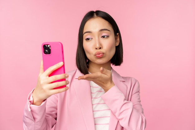 Ragazza asiatica alla moda della donna di affari in vestito che prende selfie sulla chat video dello smartphone con l'app del telefono cellulare che posa contro lo sfondo rosa dello studio
