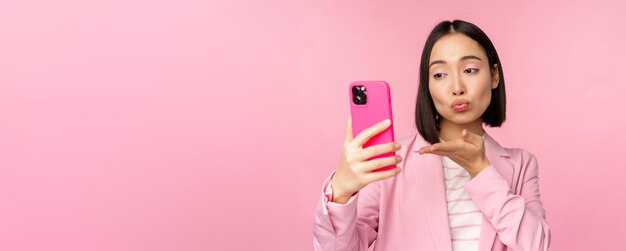 Ragazza asiatica alla moda della donna di affari in vestito che prende selfie sulla chat video dello smartphone con l'app del telefono cellulare che posa contro lo sfondo rosa dello studio