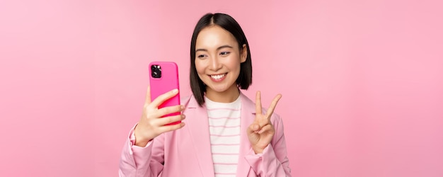 Ragazza asiatica alla moda della donna di affari in vestito che prende selfie sulla chat video dello smartphone con l'app del telefono cellulare che posa contro lo sfondo rosa dello studio