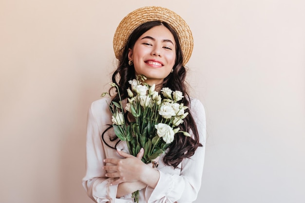 Ragazza asiatica allegra in cappello di paglia che tiene fiori Giovane donna con eustomi bianchi che esprimono felicità