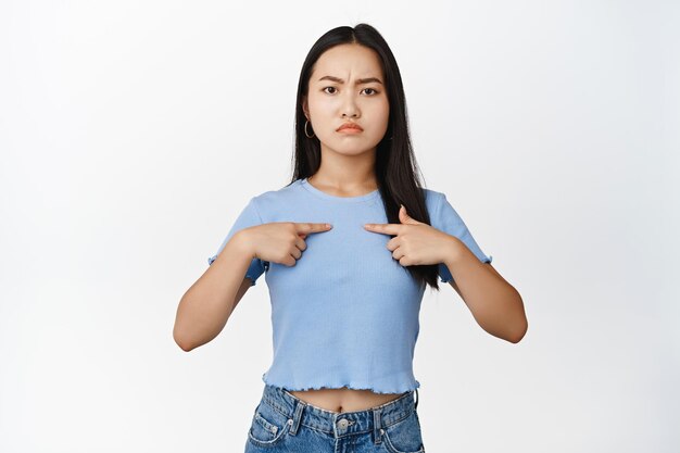 Ragazza asiatica arrabbiata che punta le dita contro se stessa accigliata e imbronciata sconvolto in piedi in maglietta blu su sfondo bianco