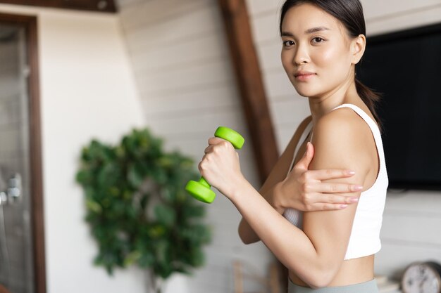 Ragazza asiatica attiva e sana con un corpo in forma che fa esercizi di fitness a casa sollevando manubri e...