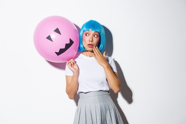 Ragazza asiatica carina e sciocca in parrucca blu, che celebra halloween, guardando sorpreso il palloncino con la faccia spaventosa.