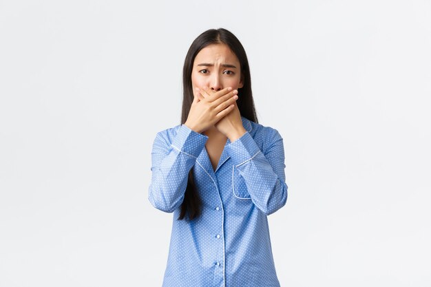 Ragazza asiatica carina preoccupata in pigiama blu accigliata e sembra preoccupata mentre chiude la bocca con le mani, copre le labbra, reagendo a notizie terribili, in piedi sfondo bianco sconvolto.