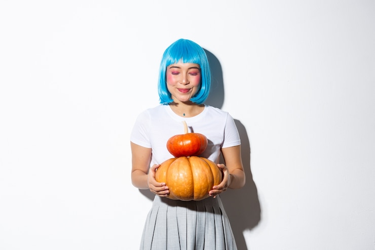 Foto gratuito ragazza asiatica carina sognante in piedi con due zucche in parrucca blu e costume di halloween, sorridente con gli occhi chiusi.