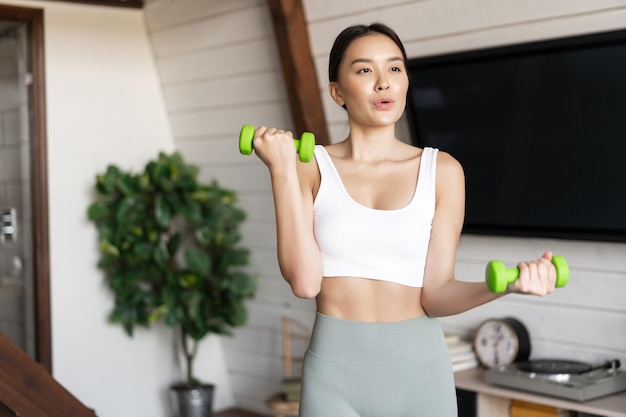 Ragazza asiatica di forma fisica che fa allenamento a casa in soggiorno sollevando manubri indossando abbigliamento attivo per spo...