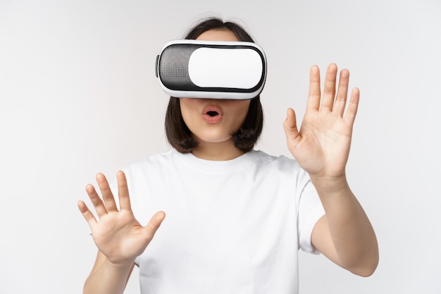 Ragazza asiatica divertita che usa le cuffie per realtà virtuale con occhiali VR e raggiunge le mani nello spazio vuoto toccando smth aumentato in piedi su sfondo bianco