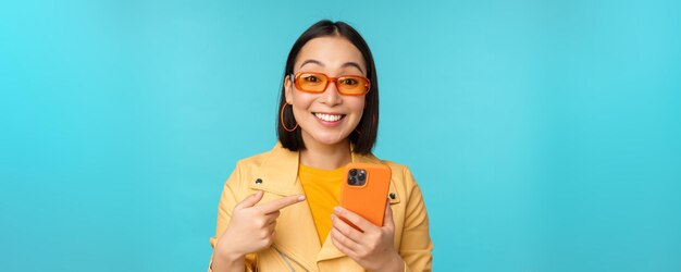 Ragazza asiatica entusiasta in occhiali da sole alla moda che punta il dito verso lo smartphone che mostra il telefono cellulare sta