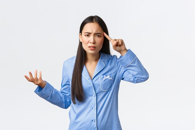 Ragazza asiatica infastidita e confusa che si lamenta per qualcuno che si comporta da stupido o pazzo, indossa un pigiama blu, alza la mano frustrata e fa rotolare il dito sulla tempia con sgomento, rimproverando qualcuno