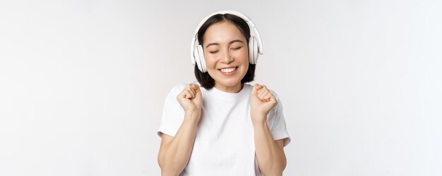 Ragazza asiatica moderna che balla musica d'ascolto con le cuffie che sorridono felice in piedi in maglietta sopra priorità bassa bianca