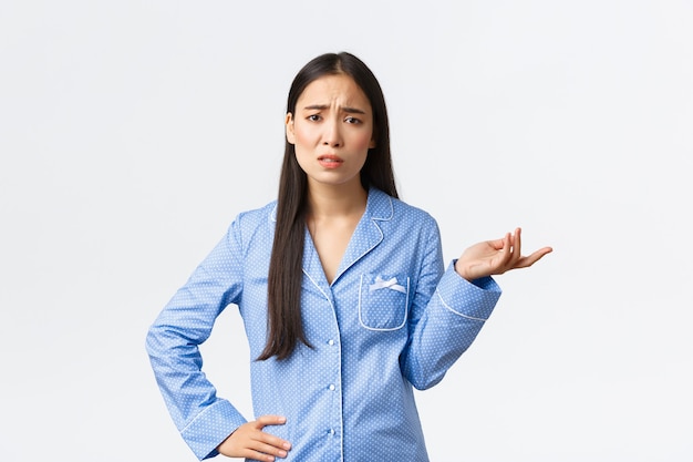 Ragazza asiatica scettica e frustrata in pigiama blu che si lamenta, discute di qualcosa, si acciglia e alza la mano perplessa, non riesce a capire cosa sta succedendo, sembra confusa.