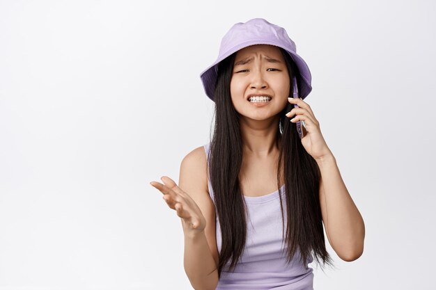 Ragazza asiatica sconvolta che sembra turbata mentre ha una telefonata accigliata e fissa angosciata avendo sconvolgente conversazione su sfondo bianco