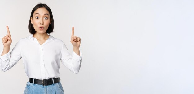 Ragazza asiatica sorpresa che punta il dito verso l'alto mostrando informazioni banner o pubblicità in piedi su sfondo bianco in camicetta Spazio di copia