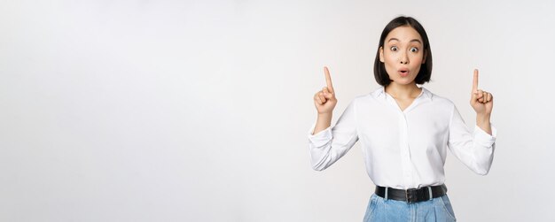 Ragazza asiatica sorpresa che punta il dito verso l'alto mostrando informazioni banner o pubblicità in piedi su sfondo bianco in camicetta Spazio di copia