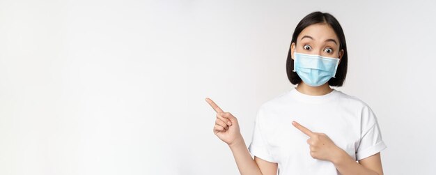 Ragazza asiatica sorpresa in maschera medica che punta le dita a sinistra mostrando un'offerta promozionale alzando le sopracciglia amaz