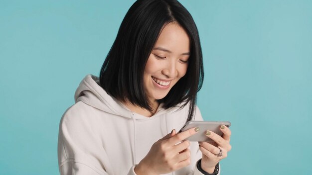 Ragazza asiatica sorridente che gioca emotivamente ai giochi sullo smartphone che partecipa alla competizione su sfondo colorato