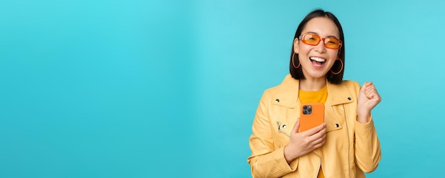 Ragazza asiatica sorridente entusiasta in occhiali da sole che tiene il telefono cellulare e balla ridendo con lo smartphone in piedi su sfondo blu