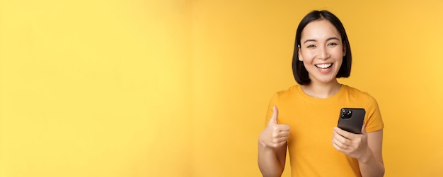 Ragazza asiatica sorridente felice che tiene il telefono cellulare e che mostra i pollici in su raccomandando l'applicazione su smar