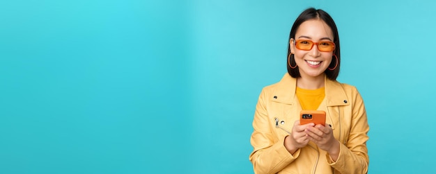 Ragazza asiatica sorridente in occhiali da sole utilizzando l'app per smartphone che tiene il telefono cellulare in piedi su sfondo blu