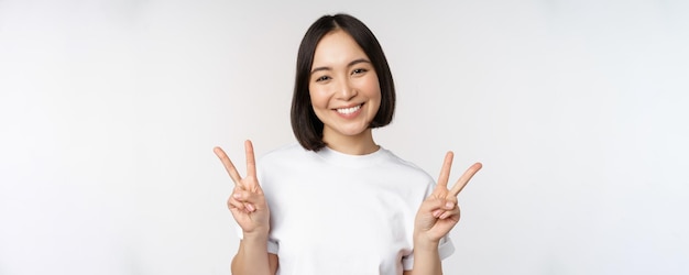Ragazza asiatica sveglia che mostra il segno di pace che sorride e che guarda felice alla macchina fotografica che indossa lo sfondo bianco dello studio della maglietta