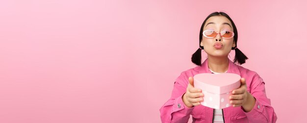 Ragazza asiatica sveglia che ti dà un regalo in una scatola a forma di cuore che bacia e che sorride concetto di vacanza e celebrazione in piedi su sfondo rosa