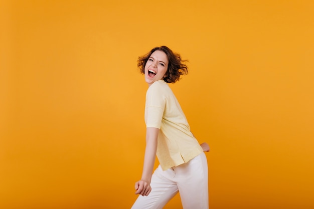 Ragazza attiva con taglio di capelli alla moda che balla sulla parete arancione brillante. Affascinante signora riccia in maglietta giallo chiaro che si diverte al coperto.