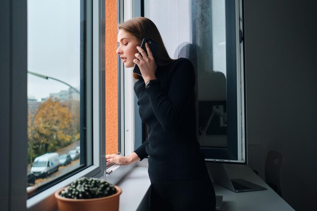 Ragazza attraente che parla al cellulare mentre guarda premurosamente fuori dalla finestra a casa