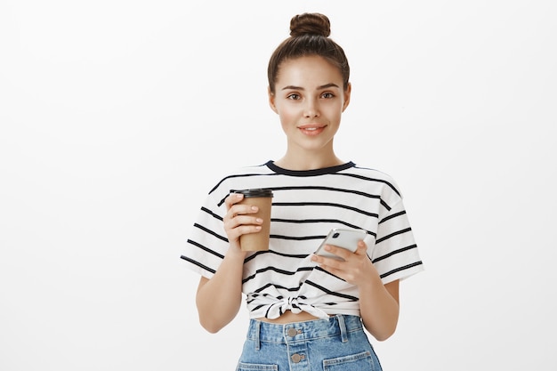 Ragazza attraente che utilizza smartphone mentre beve il caffè, che tiene tazza e telefono cellulare