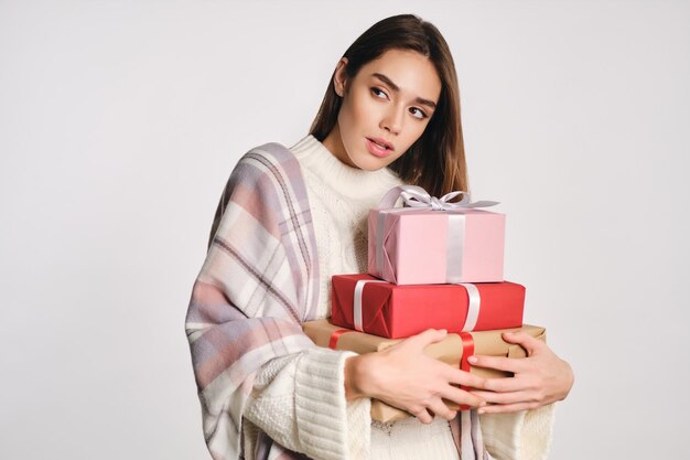 Ragazza attraente in maglione accogliente con plaid sulle spalle che tiene scatole regalo pensieroso distogliendo lo sguardo su sfondo bianco