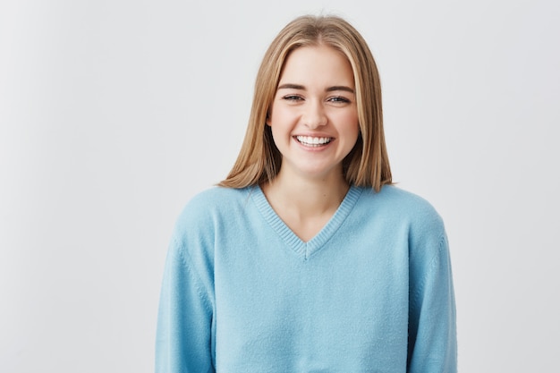 Ragazza attraente spensierata con capelli biondi, occhi scuri che indossa maglione blu sciolto che sorridono felicemente nella macchina fotografica che mostra i suoi denti bianchi mentre posa contro il muro di cemento dello studio, ridendo allo scherzo