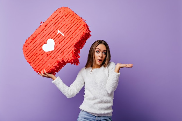 Ragazza ben vestita sorpresa che posa con la bandiera rossa. Ritratto dell'interno del blogger femminile emotivo in soffice maglione.