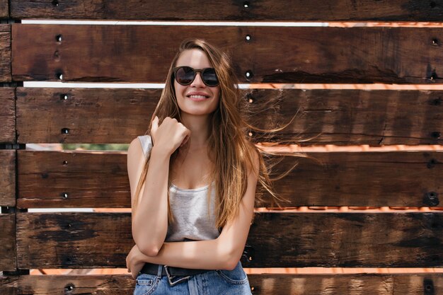 Ragazza bianca che esprime buone emozioni sulla parete di legno. Ritratto di magnifica giovane donna pallida indossa occhiali da sole scuri.