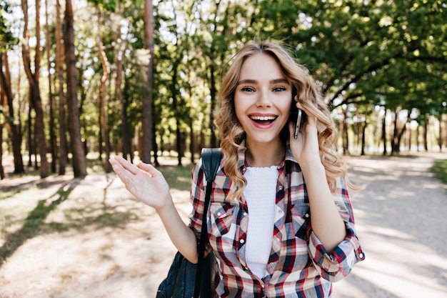 Ragazza bianca estatica parla al telefono mentre riposa nella foresta. Foto all'aperto di donna allegra con capelli ondulati in giro nel parco.