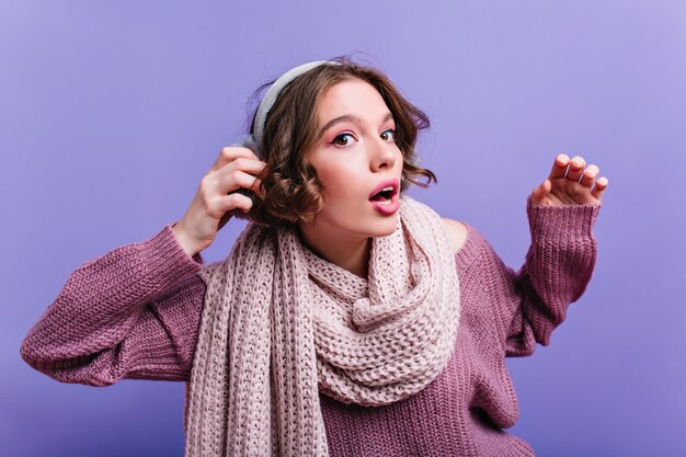 Ragazza bianca sorpresa in sciarpa lavorata a maglia lunga che posa sulla parete viola. signora bruna emotiva in accessori di lana.