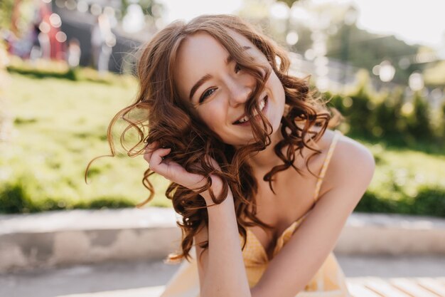 Ragazza bianca spensierata che gioca con i suoi capelli ricci mentre posa nel parco. Ritratto all'aperto di bella signora dello zenzero agghiacciante nella giornata di sole.