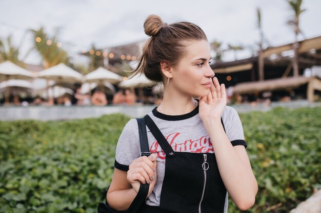 Ragazza bianca spensierata con capelli castani che distoglie lo sguardo mentre posa sulla strada. Colpo esterno di splendida donna europea in t-shirt alla moda trascorrere il fine settimana nella città di villeggiatura.